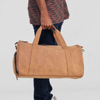 Person holding a tan leather duffel bag against a plain background
