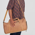Man holding a brown leather duffel bag against a plain background