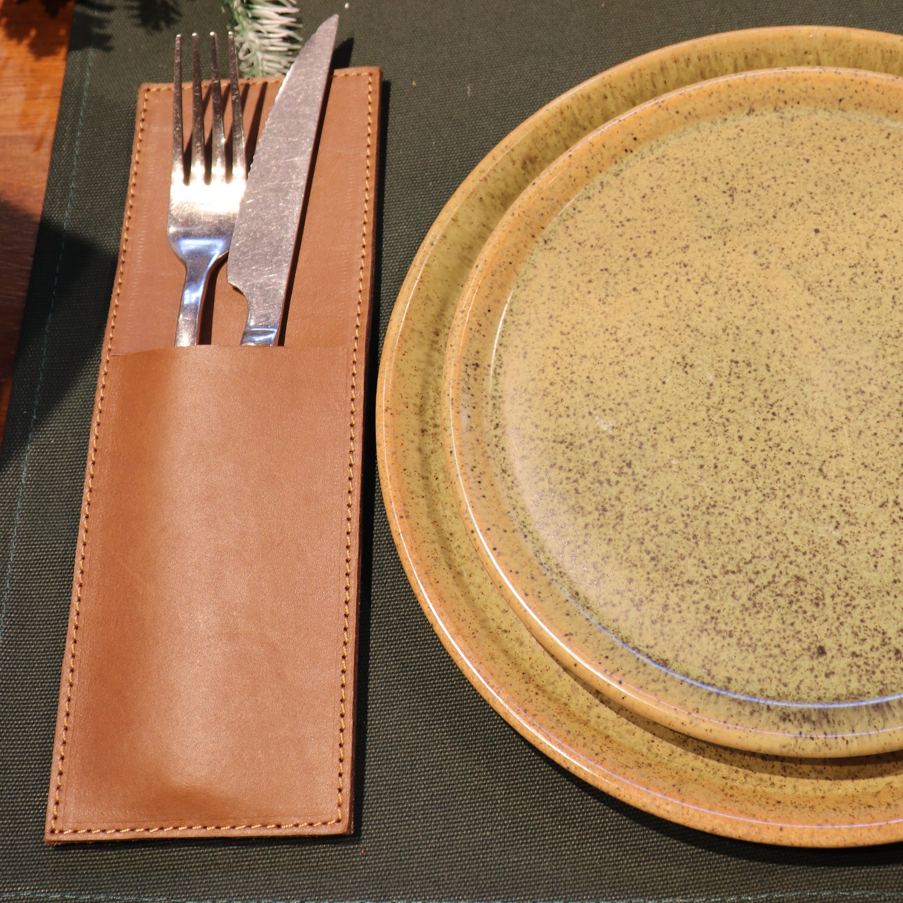 A festive tabletop setting featuring a dark olive green placemat, a speckled beige ceramic dinner plate, a matching charger plate, and a brown leather knife and fork holder with silver cutlery. The background is decorated with a pine garland and red holiday ornaments.