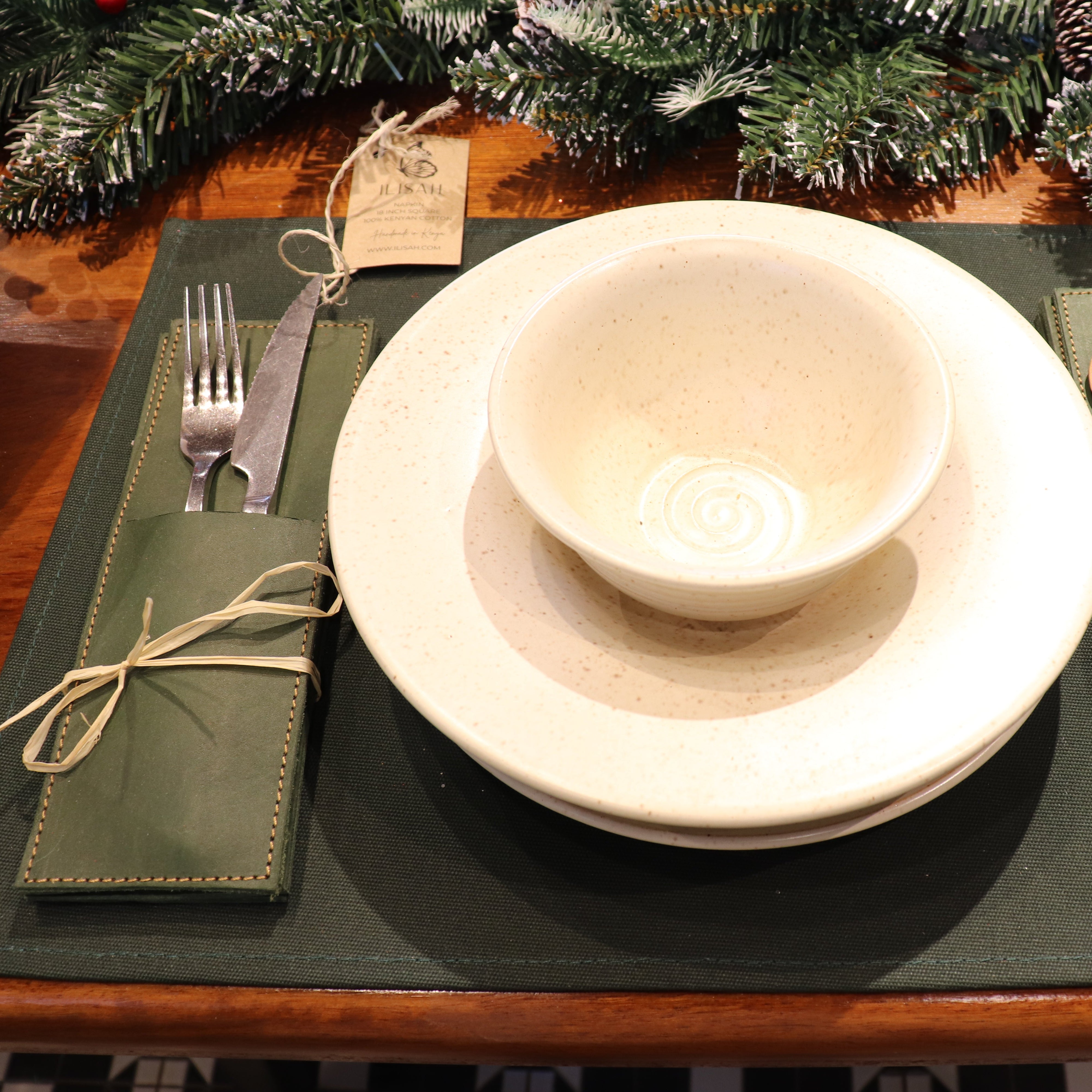 A festive table setting featuring a dark green placemat with a matching green napkin holder tied with twine, holding silver cutlery and a small wooden spoon. In the center is a stack of cream-colored ceramic plates with a matching cream bowl.