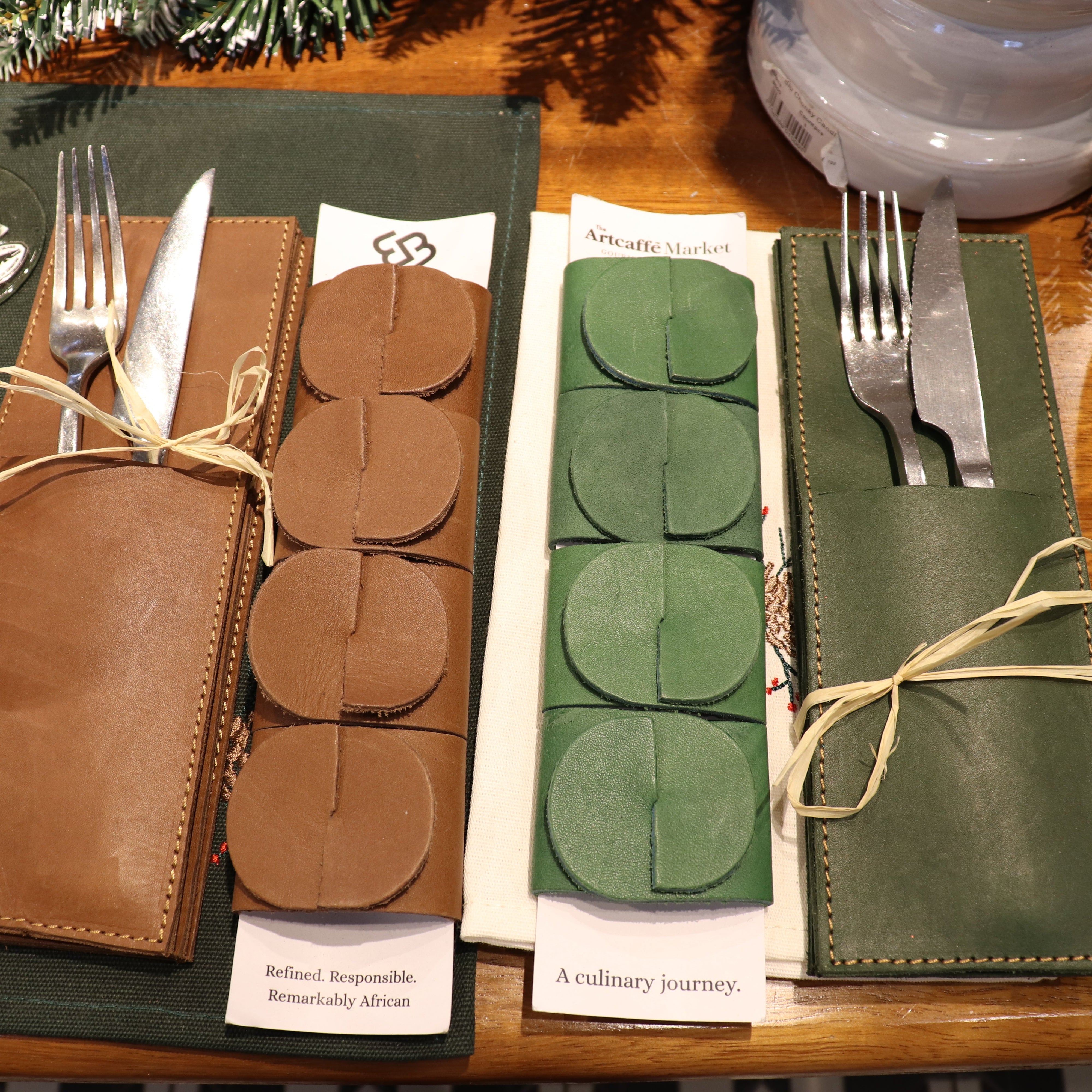 Three leather cutlery sleeves in brown, green, and white, each holding a fork and knife, arranged on a wooden table with tableware and evergreen branches around them.
