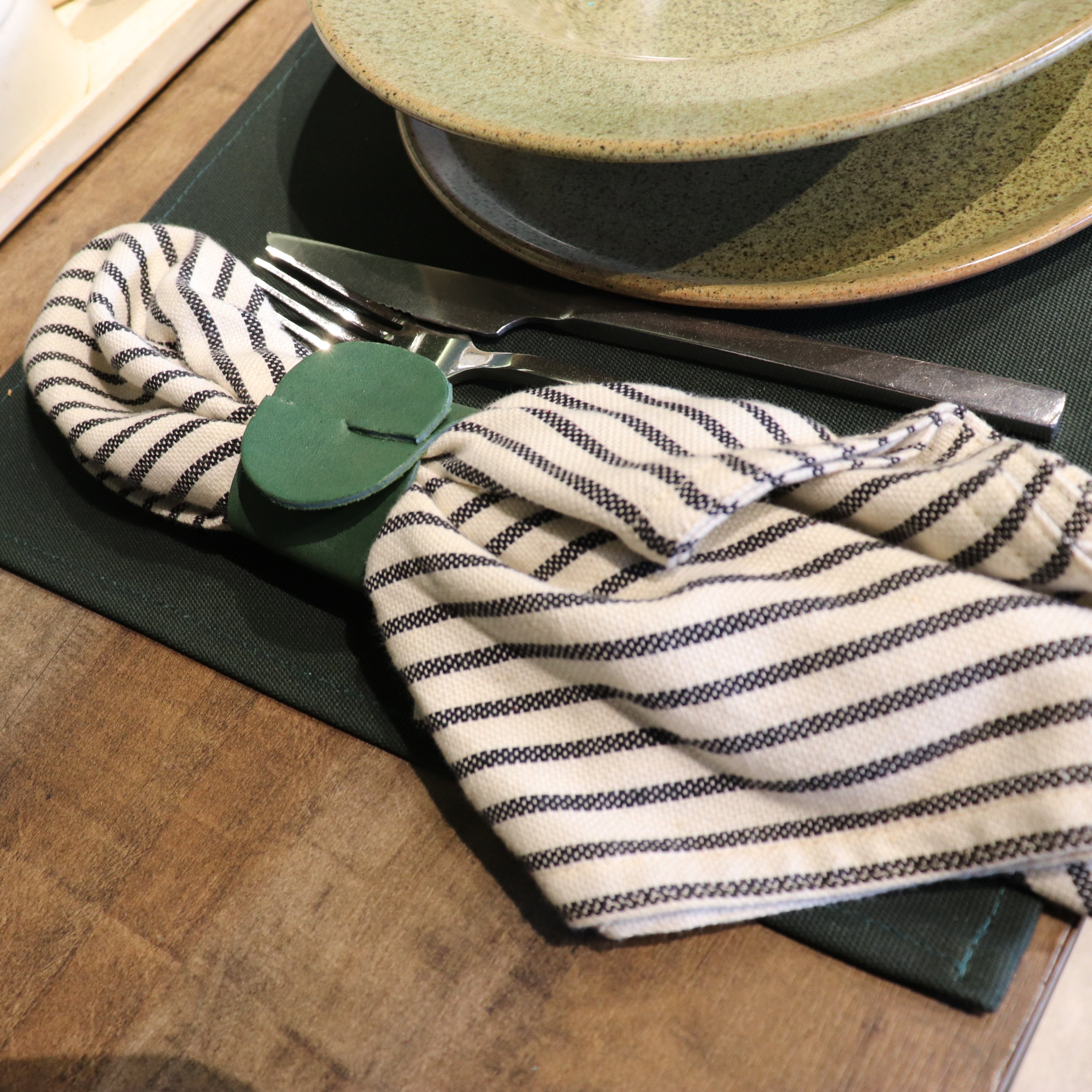 A close-up of a table setting featuring a cream and black striped cloth napkin with a green plastic napkin clip, placed on a dark green placemat next to silver flatware, with ceramic plates in the background.