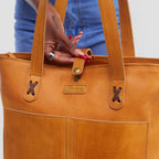 Brown leather tote bag held by a person wearing blue jeans.
