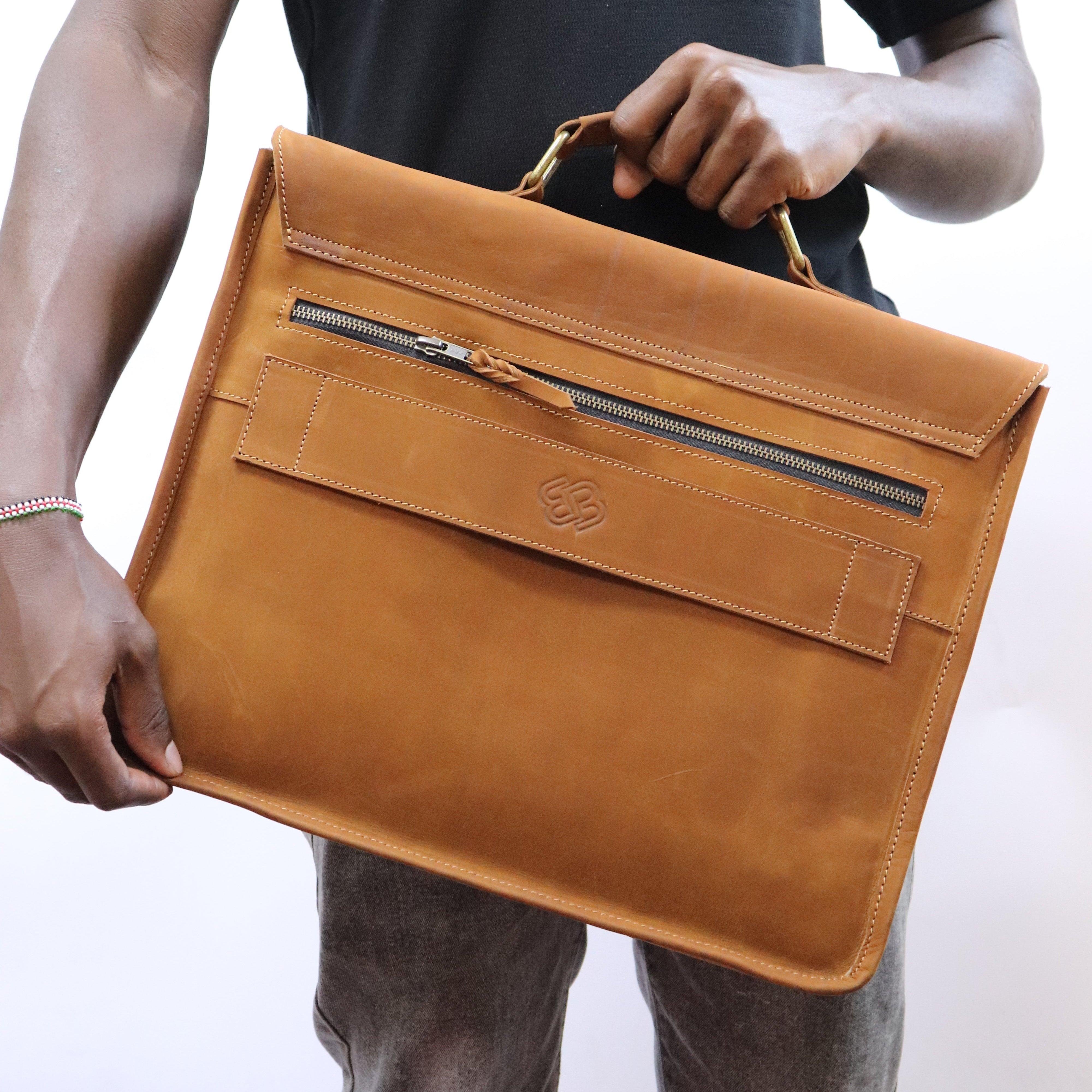 A person holding a tan leather briefcase-style bag with a top handle and front zip pocket, wearing a black t-shirt and gray jeans against a plain light background.