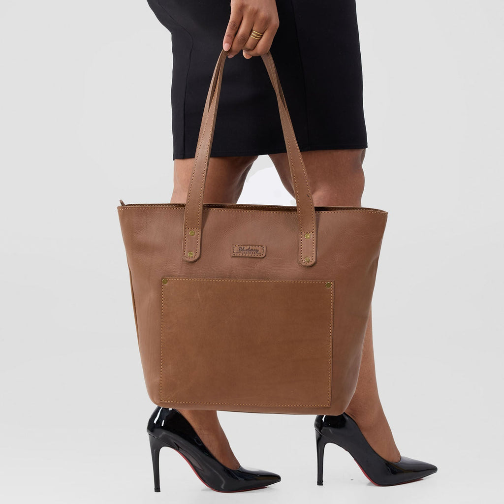 Brown tote bag held by a person wearing black high heels on a white background