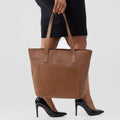 Brown tote bag held by a person wearing black high heels on a white background