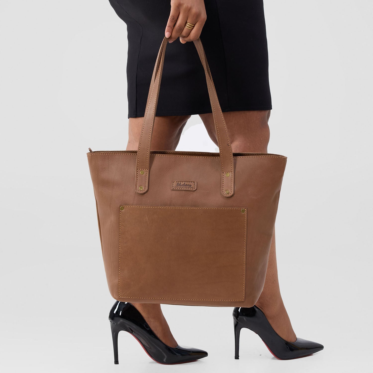 Brown tote bag held by a person wearing black high heels on a white background