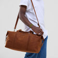 Person holding a brown leather duffel bag against a plain background