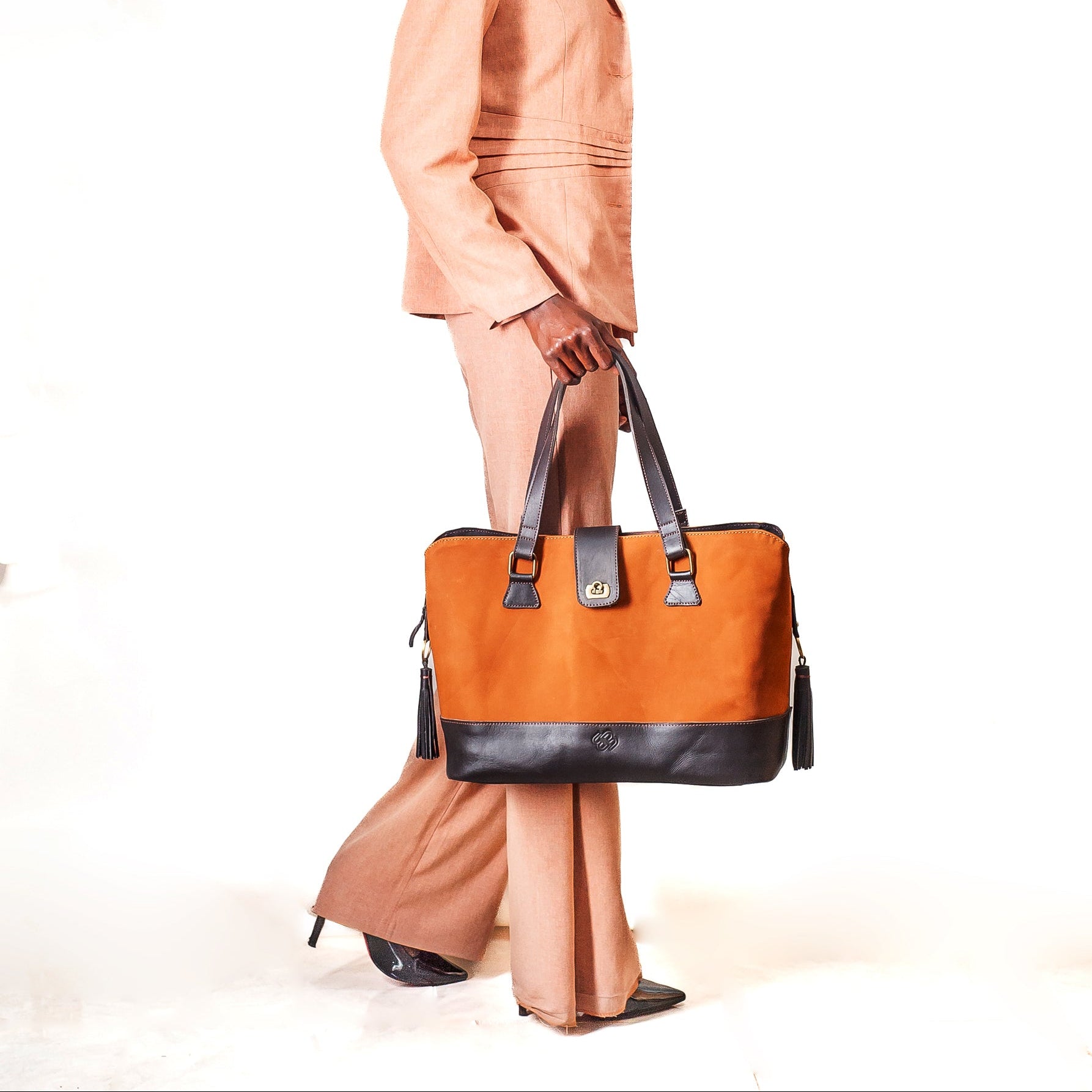 A model in a peach-colored linen suit holds a structured orange and black canvas tote bag against a plain white studio background.