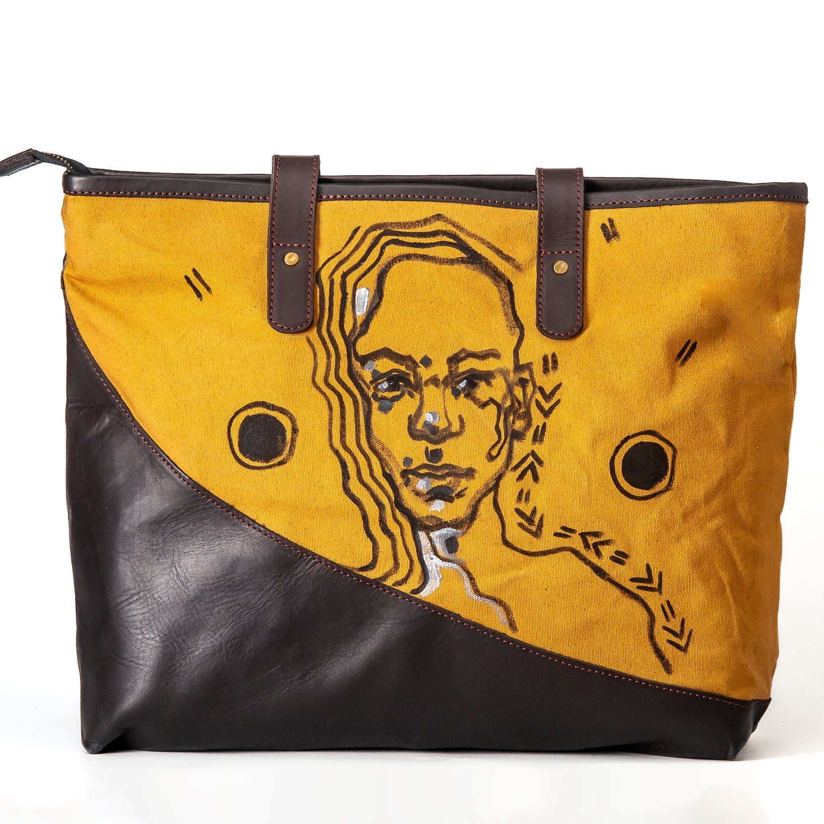 A large tote bag with a diagonal split design, featuring a mustard yellow canvas upper section with a stylized portrait illustration and black circular details, combined with a dark brown leather lower section and matching brown leather handles.