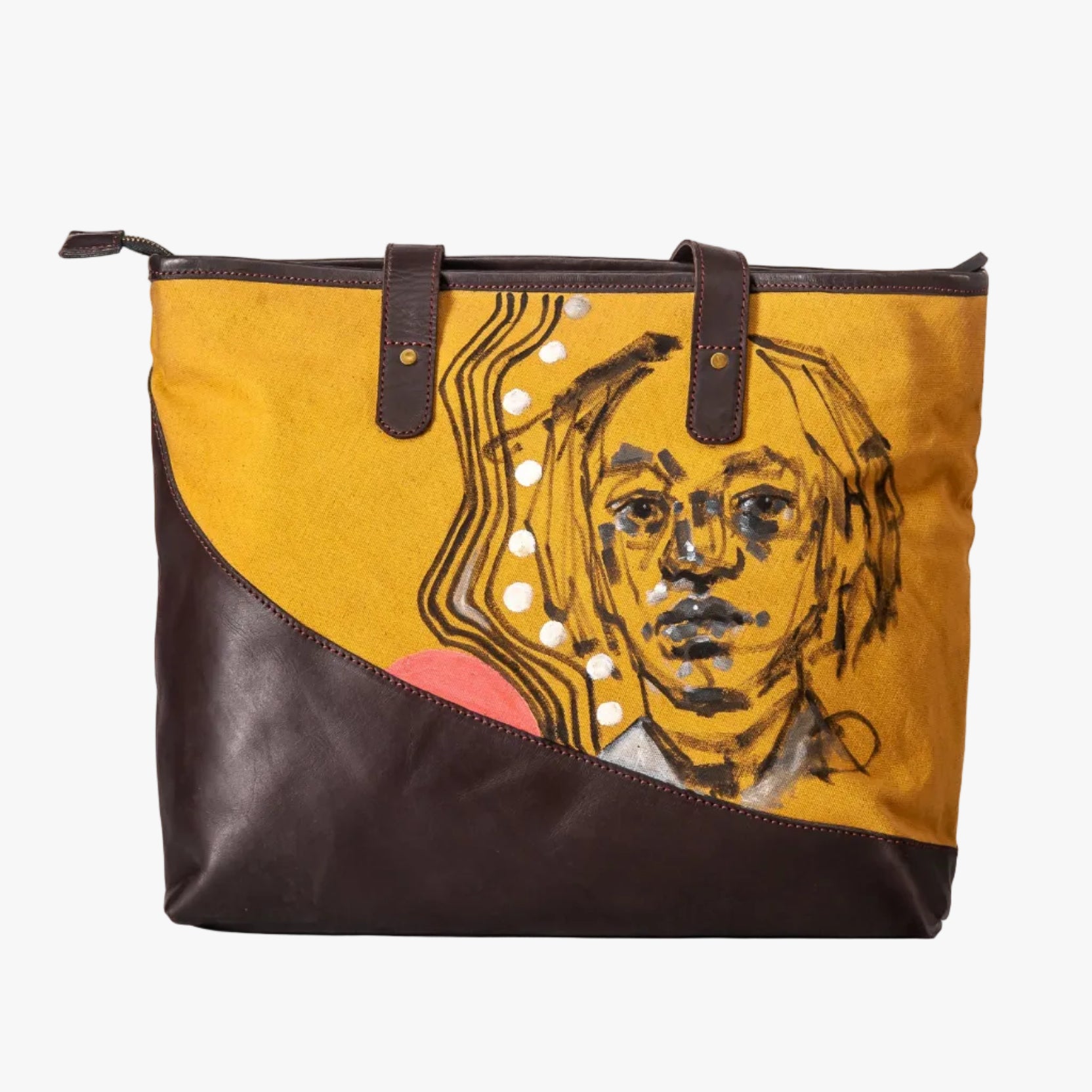 A large tote bag with a diagonal split design of mustard yellow canvas and dark brown leather. The canvas side features a graphic illustration of a face with black lines, white dots, and wavy patterns, while the leather side is solid dark brown.