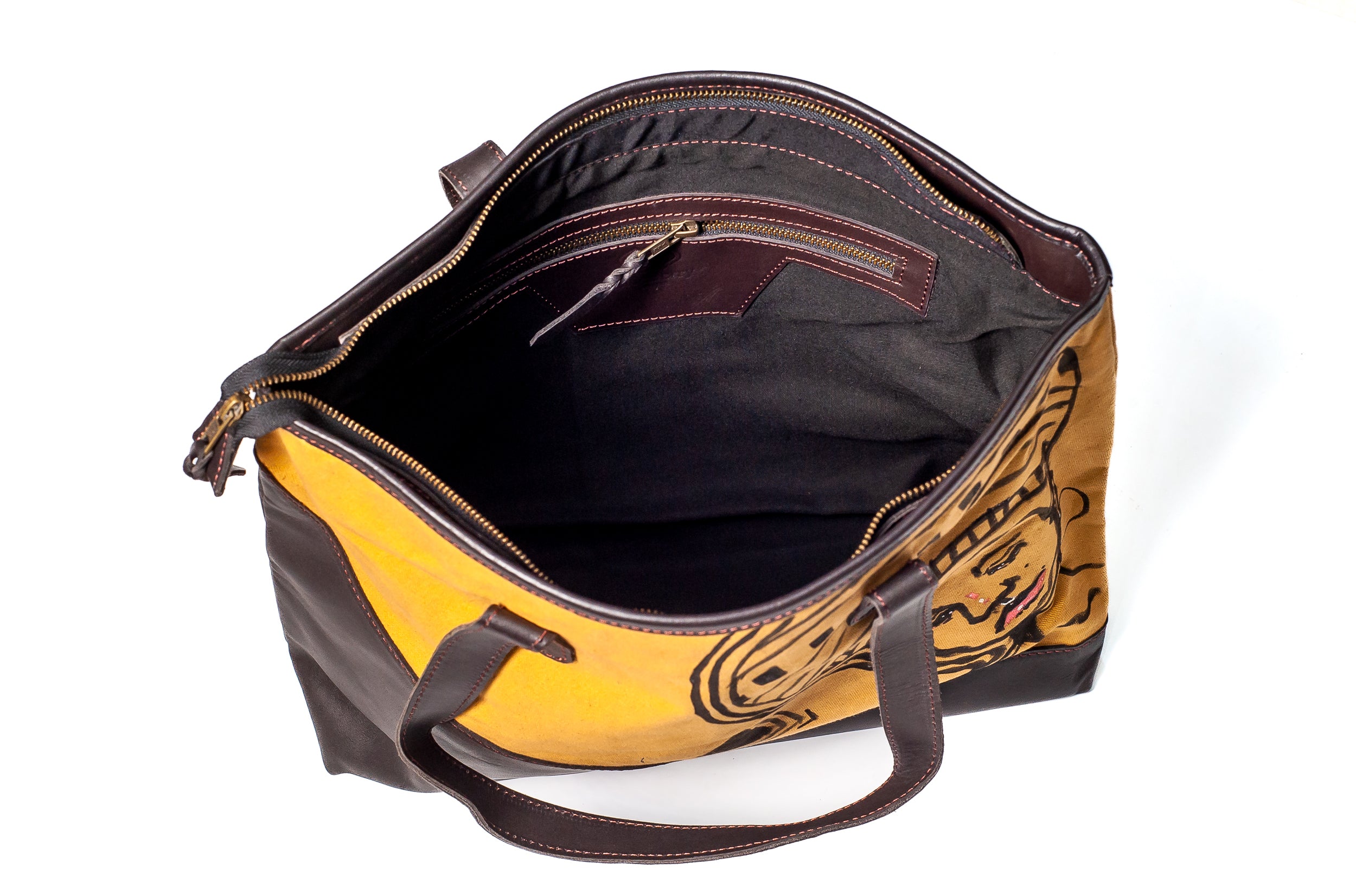 An open tote bag with a mustard yellow and black graphic patterned exterior, dark brown leather handles and trim, and a black interior lining, shown on a white background.
