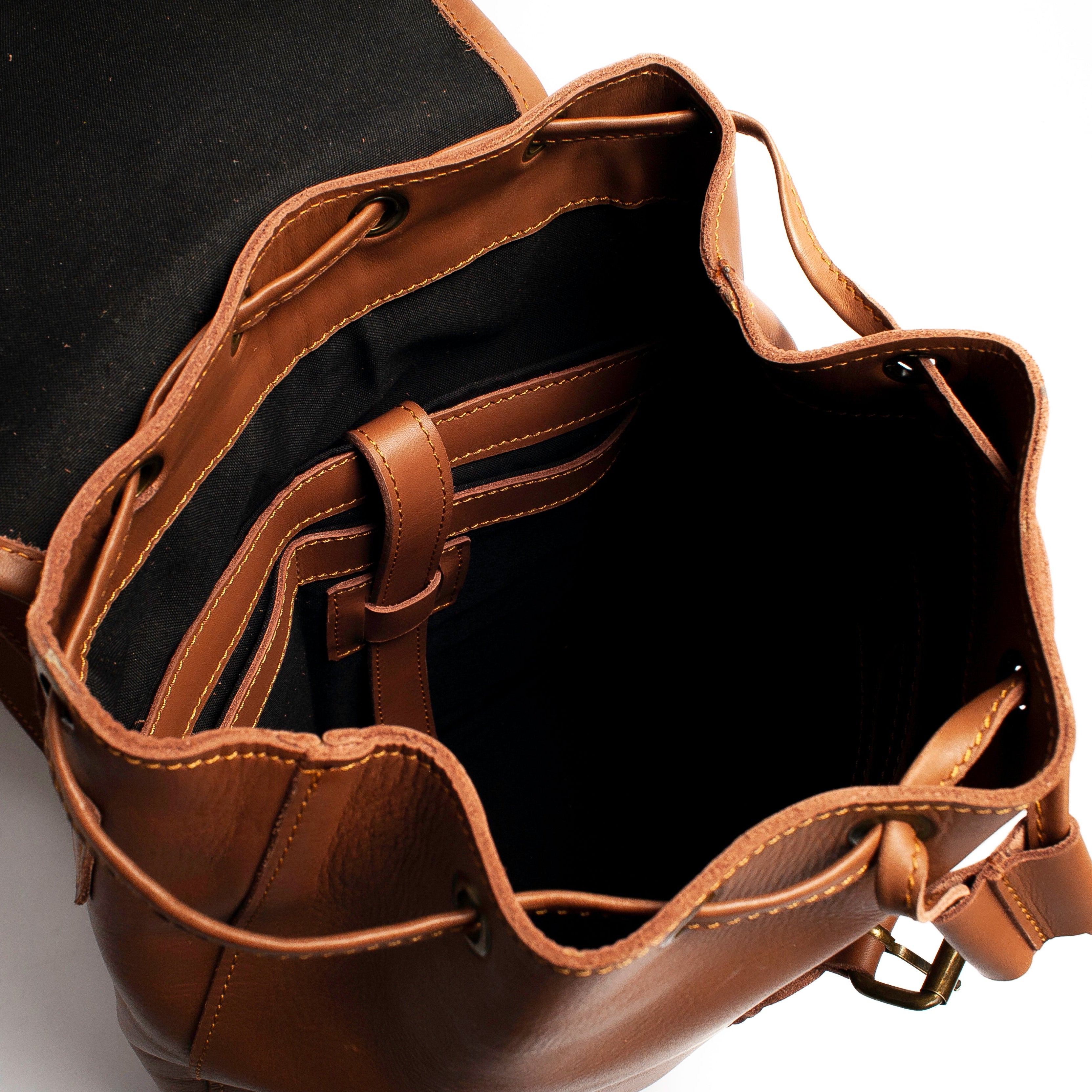 A close-up shot of a brown leather bucket bag shown open, displaying its black fabric lining, interior straps, and drawstring closure against a white background.