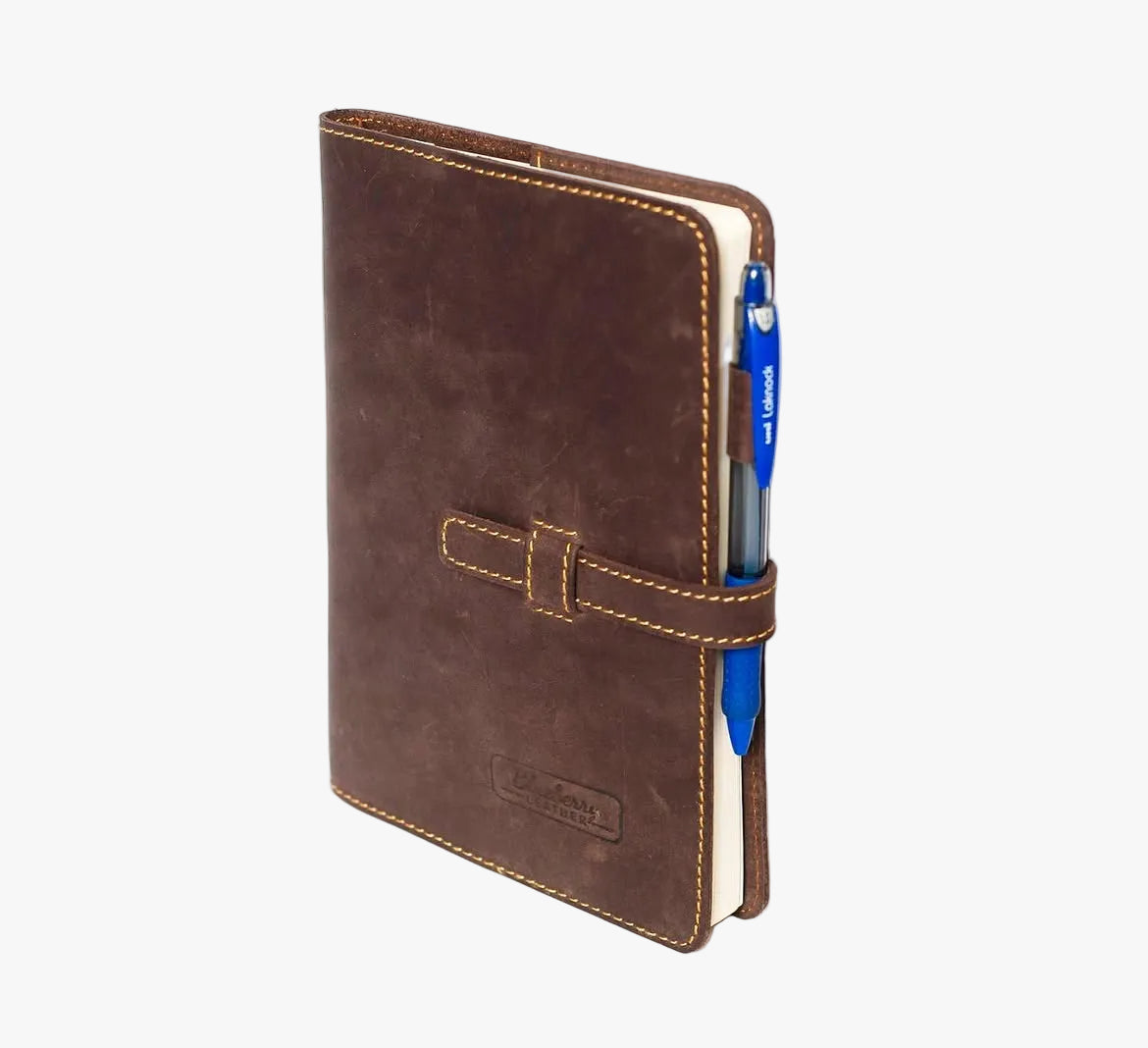 A brown leather notebook cover with a strap closure and visible contrast stitching, shown standing upright on a white background. A blue pen is tucked into the side pocket, and a small embossed brand plaque is visible near the bottom.