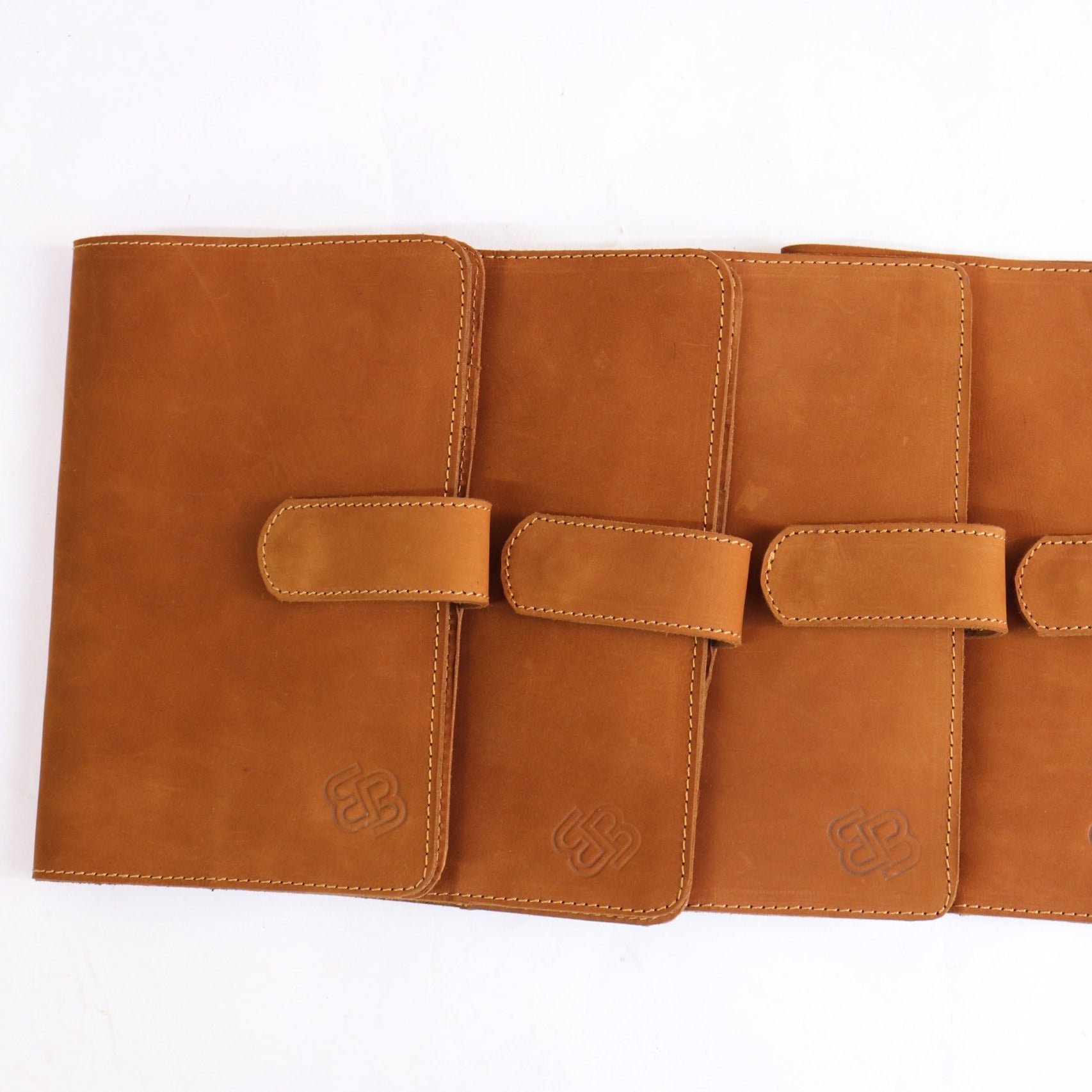Four tan leather planner covers arranged in a row on a white background, each with a strap closure and a small embossed logo near the bottom.